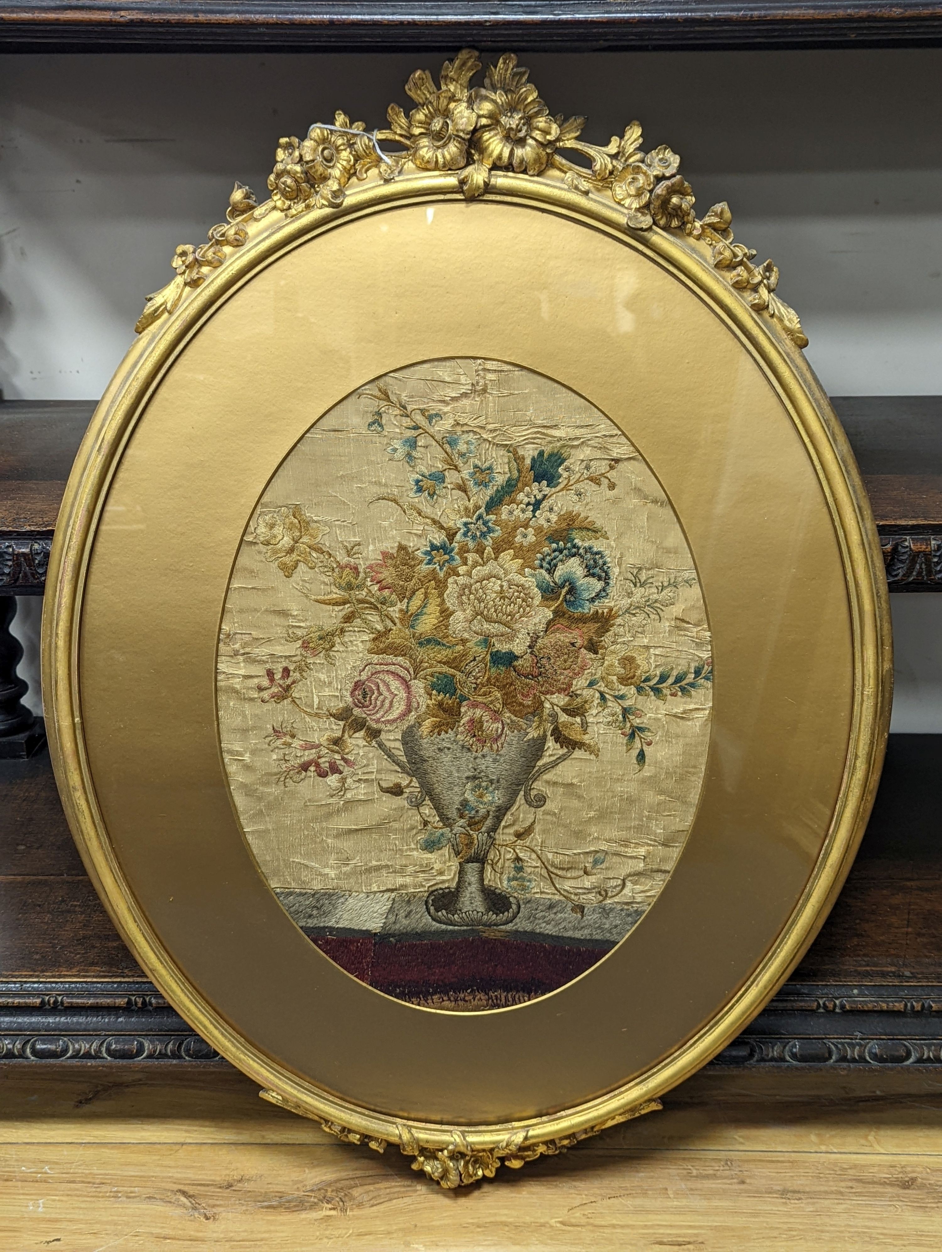 A Georgian oval silk embroidery panel depicting a two handled classical urn containing mixed flowers 46cm x 34cm, glazed gilt frame